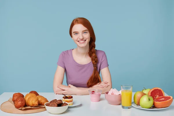 Lachende roodharige meisje met gevlochten haren zitten aan een tafel, ongeveer om te ontbijten, kijken naar de camera, geïsoleerd op blauwe muur. Op de tafel leggen bakken en vers fruit sap yoghurt. Voedsel concept — Stockfoto