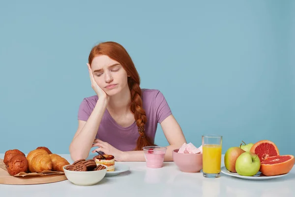 Studio shot of red-haired girl looking with discontent sadness on baking products thinks about what food to eat, fresh fruit juice yogurt on the other side of table, isolated on a blue background — Stock Photo, Image