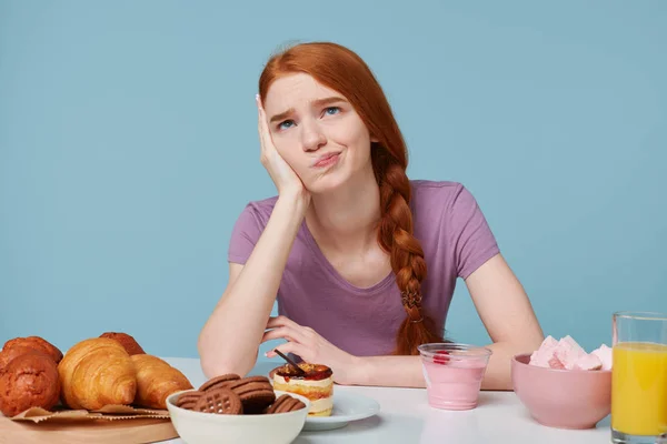 Studiofoto eines traurigen rothaarigen Mädchens, Zweifel grübeln über Essen, Gesundheit, Ernährung, zusätzliche Kalorien, Backwaren und frischer Fruchtsaftjoghurt liegen auf dem Tisch, isoliert auf blauem Hintergrund — Stockfoto