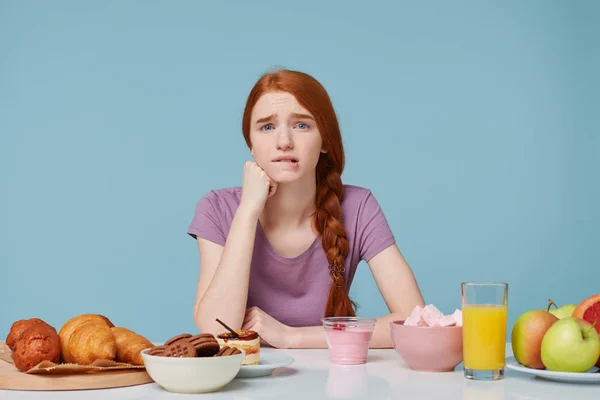 Sad red-haired girl looking camera bites lip, worries doubts about nutrition, health, thinks about diet, extra calories, baking food and fresh fruit juice yogurt lay on the table, on a blue background — Stock Photo, Image