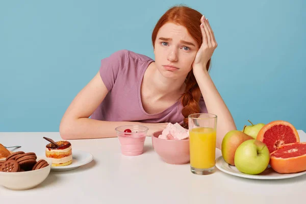Menina ruiva jovem senta-se à mesa com a cabeça na mão, infelizmente, entende a importância da fruta fresca e comida saudável, mas quer comer bolo, isolado em um fundo azul. — Fotografia de Stock
