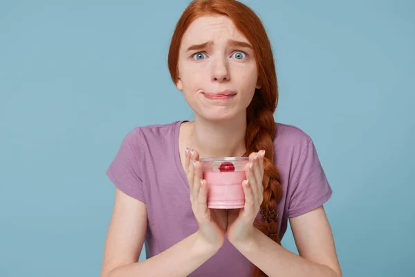 Chica pelirroja sostiene yogur de frutas en las manos, se ve preocupada, se muerde el labio, mira la cámara con una mirada compasiva, se preocupa por lo que come, piensa en qué tipo de dieta seguir, sobre un fondo azul — Foto de Stock