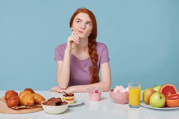 Foto eines rothaarigen Mädchens, das mit Unzufriedenheit nach oben blickt, Zweifel an Ernährung, Gesundheit, Ernährung, zusätzlichen Kalorien, Backwaren und frischem Fruchtsaftjoghurt liegen auf dem Tisch, auf blauem Hintergrund — Stockfoto