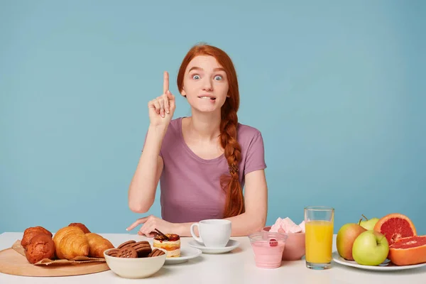 A lovely girl is sitting at a table during lunch time, a good idea suddenly came to mind,raised index finger. On the table is pastries and fresh, healthy products, fruits,isolated on a blue background — Stock Photo, Image