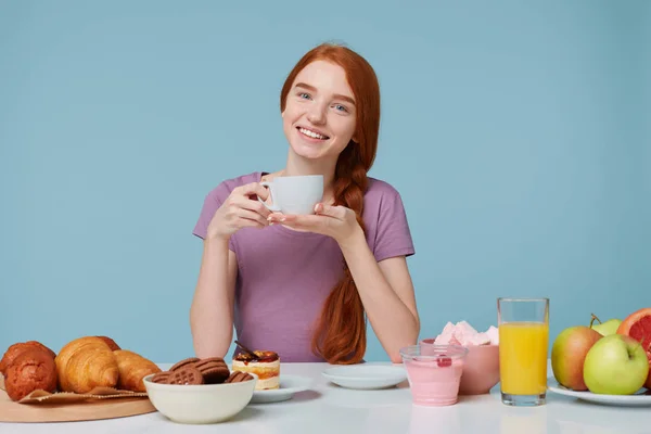 Chica pelirroja sentada en una mesa con la cabeza ligeramente inclinada, sonrisas sostiene taza blanca con deliciosa bebida en las manos, tiene el almuerzo mirando cámara. En la mesa de productos para hornear y alimentos frescos ponen — Foto de Stock