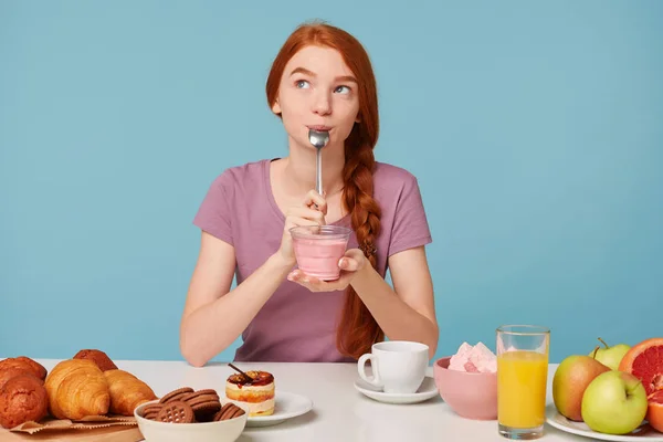 Nice red-haired girl with a braid sits at a table, has lunch, holds in her hands a glass, dreamingly looks at left upper corner and licks teaspoon with pleasure after tasting yummy yogurt. — Stock Photo, Image