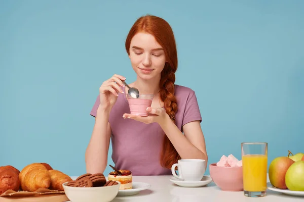 Una linda pelirroja con una trenza se sienta en una mesa, almuerza, tiene en sus manos un yogur de cereza, lo mira dentro de la tentación y va a comer con una cucharadita . — Foto de Stock