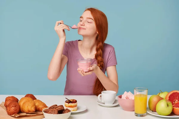 Cute red-haired girl trying tasting cherry yogurt, closed her eyes from pleasure, licks a teaspoon sitting at the table during lunch, pastries on the table and fresh fruit, against a blue background. — Stock Photo, Image