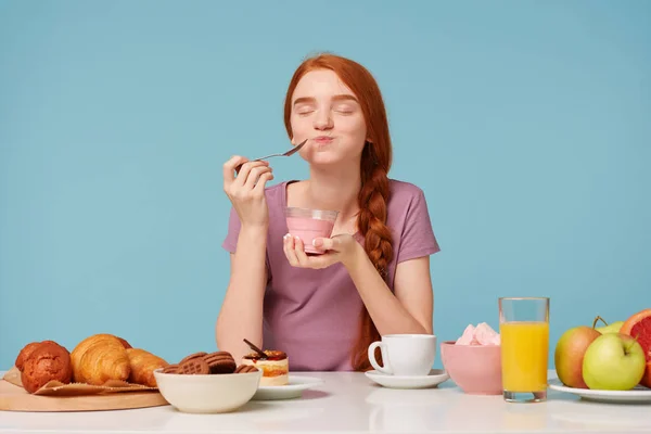 Nice red-haired girl trying tasting cherry yogurt wirh a teaspoon, closed her eyes from pleasure, sitting at the table during lunch, pastries on the table and fresh fruit, against a blue background. — Stock Photo, Image