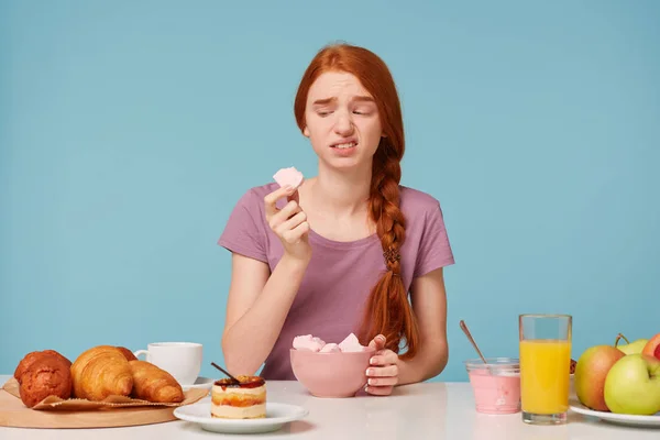 Ein rothaariges Mädchen, das während des Frühstücks an einem Tisch sitzt, probiert einen Fruchtmarshmallow, hält ein Stück in der Hand und schaut ihn angewidert an, ihr gefiel der Geschmack auf blauem Hintergrund nicht. — Stockfoto