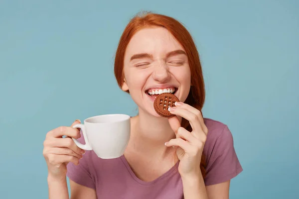 Close up of a lovely beautiful red-haired girl with closed eyes, feels pleasure, keeps in hand white cup with drink, with appetite bites yummy chocolate cookie, over blue background — стоковое фото