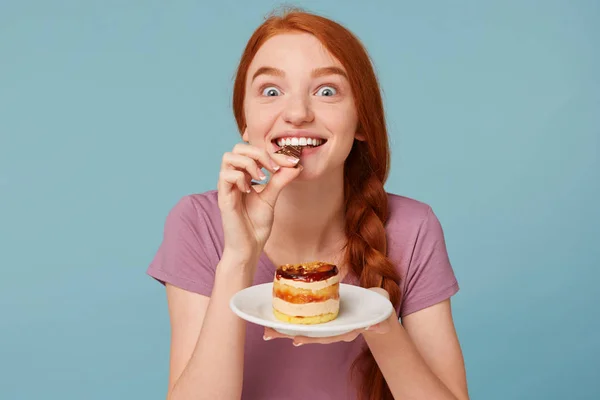 Nahaufnahme eines rothaarigen Mädchens, das eifrig einen leckeren Kuchen probiert, ein Stück Schokolade abbeißt und auf blauem Hintergrund einen Teller in der Hand hält. — Stockfoto