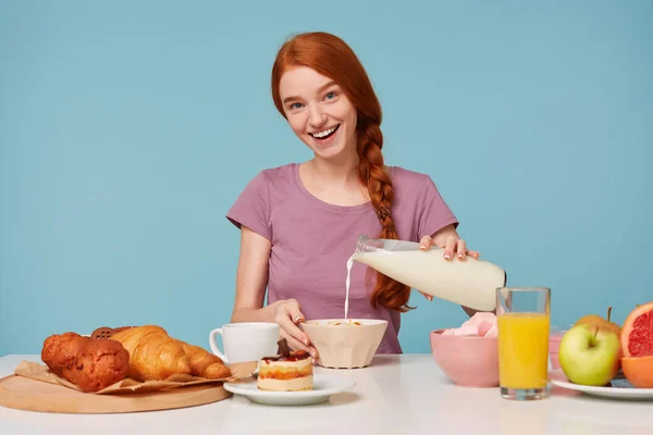 Menina alegre tipo alegre ruiva sentada à mesa vai tomar café da manhã. Despeje leite em um prato com mexilhões. Sorrindo afetuosamente, olhando para a câmera. Isolado sobre um fundo azul . — Fotografia de Stock