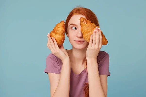 Brincalhão alegre menina ruiva segurando dois croissants em suas mãos, olha para longe cobre fecha o olho com um croissant, isolado em um fundo azul . — Fotografia de Stock