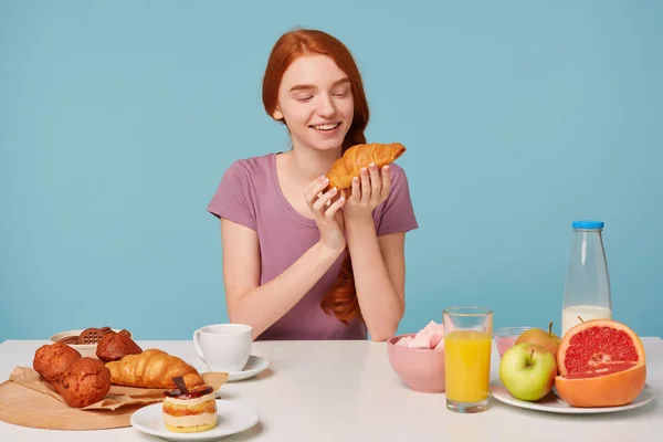 Leuk roodharige meisje met een vlecht zit aan een tafel, Ontbijt, met opwinding genoegen kijkt croissants, gebak op tafel en vers fruit, heeft op een blauwe achtergrond — Stockfoto