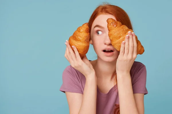 Close up de surpreendido e surpreendido menina ruiva isolado em um fundo azul segurando dois croissants em suas mãos, fecha-se o olho olhando para a esquerda no espaço de cópia vazia para publicidade . — Fotografia de Stock