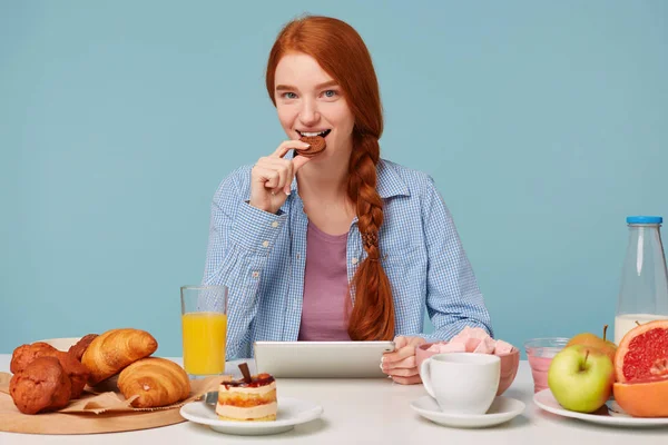 Nettes attraktives rothaariges Mädchen mit einem Lächeln, das an einem Tisch sitzt und Kekse isst, in ihren Händen ein Tablet, auf dem die Nachrichten gelesen werden, in die Kamera blickt und vor blauem Hintergrund lächelt. — Stockfoto