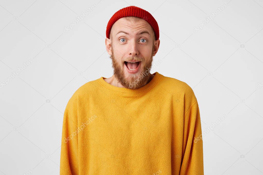 Glad ginger-breaded male with happy amazed expression, wears red hat and casual sweater, rejoices successfully made project, stares and poses alone against white background. People, emotions,lifestyle