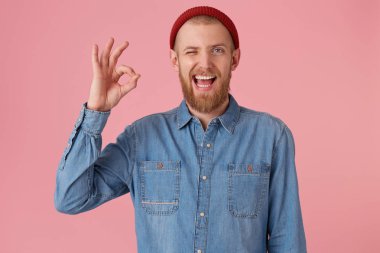 Tamam işareti, çekici sakallı erkek moda kot gömlek kırmızı şapka, memnun ifade ile gösterir geliyor toplantıdan sonra memnun izole pembe arka plan üzerinde. İnsan yüz ifadeleri, beden dili