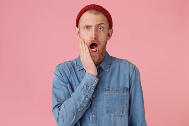 Hayret endişeli, açılan sürpriz ve kırmızı kalın sakallı bırakılan çene, kırmızı şapkalı adam elini yüzünü yakınındaki tutar ağzına moda kot gömlek, pembe arka plan izole giyer