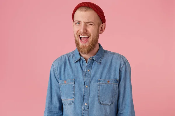Positive bearded male with fashioned denim shirt red hat, has playful expression, winks, mouth opened, encourages supports, isolated over pink background. Human facial expressions, body language — Stock Photo, Image