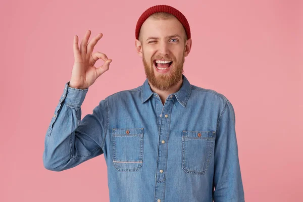 Attractive bearded male with fashioned denim shirt red hat, satisfied expression, shows okay sign, feels glad after meeting, isolated over pink background. Human facial expressions, body language — Stock Photo, Image