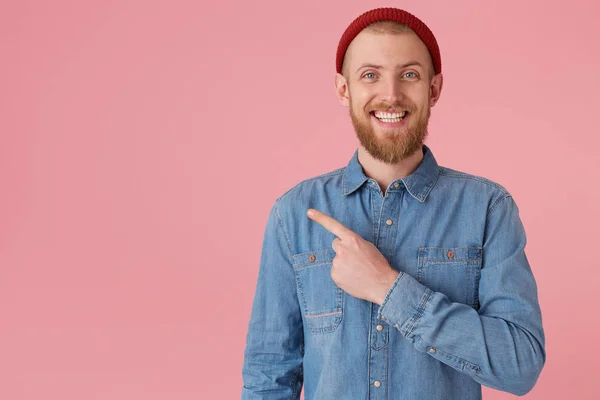 Joyfully smiling blue-eyed guy in red hat with red thick beard feels happiness showing white healthy teeth, in denim shirt, isolated on pink wall points with index finger to the left on copy space — Stock Photo, Image