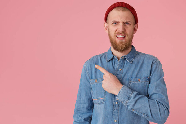Disgruntled man with a wrinkled face, makes a grimace of disgust, one corner of the lips is raised, expresses discontent, irritation,points with index finger to the left on copy space, on pink wall
