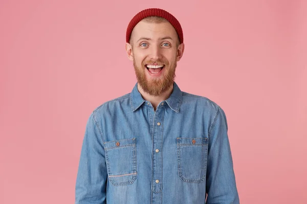 Portrait of laughing guy in red hat wears fashionable denim shirt, with red thick beard looks happy joyful, has fun, isolated on pink background — Stock Photo, Image