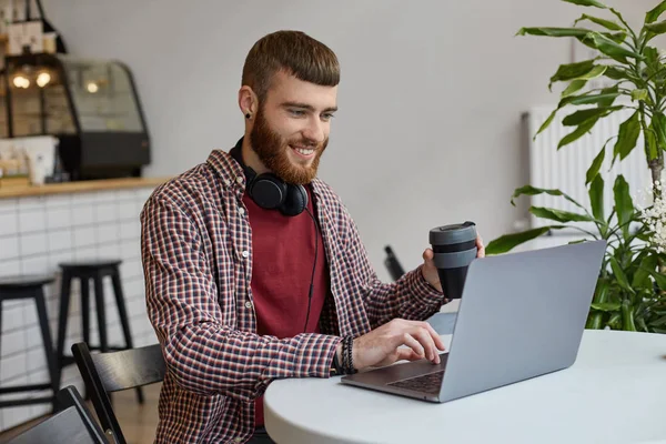 Happy young attractive ginger bearded man working at a laptop wh