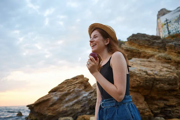 Young laughing ginger freckles lady on the beach, wears hat, eat