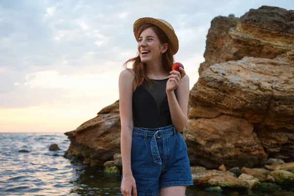 Photo of young laughing ginger freckles woman in hat, eating a p