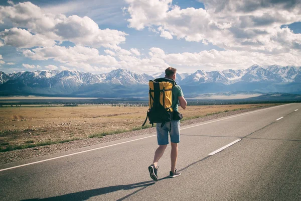 L'uomo cammina lungo una strada asfaltata con uno zaino sulla schiena che domina le montagne Foto Stock Royalty Free