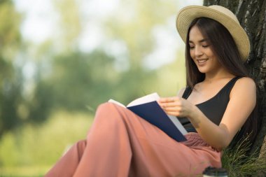 Gülümseyen mutlu genç kadın bir kitap okuyor. Kız parkta dinleniyor. Kopyala