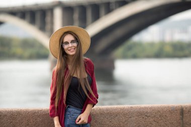Şehirde duran şapkalı ve gözlüklü mutlu kadın. Arkaplan ve köprü. Modern moda giyim tarzı. Kopyala