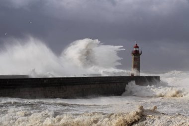 Fırtınalı karanlık bulutlu gökyüzüne karşı büyük beyaz dalgalar ve deniz feneri. Douro nehri ağzı, Porto, Portekiz.