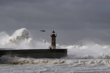 Fırtınalı karanlık bulutlu gökyüzüne karşı büyük beyaz dalgalar ve deniz feneri. Douro nehri ağzı, Porto, Portekiz.