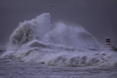 Fırtınalı karanlık bulutlu gökyüzüne karşı büyük beyaz dalgalar ve deniz feneri. Douro nehri ağzı, Porto, Portekiz.