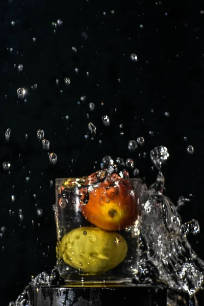 Kırmızı domates ve sarı limon, siyah arkaplanlı bir bardak suya atlıyor.