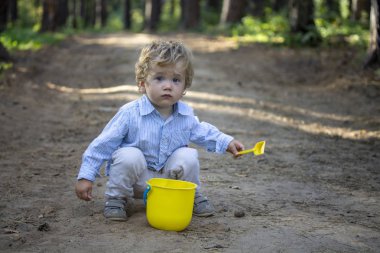 Elinde kürek ve kovayla yolda oturan küçük bir çocuk.