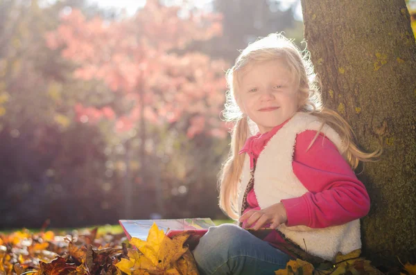 Entzückendes blondes Kind lächelt mit Buch in der Hand in herbstlicher Sonnenuntergangsatmosphäre — Stockfoto