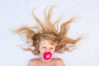 Sağlıklı ve güçlü, uzun sarı saçları ve ağzında kırmızı gül çiçeğiyle gülümseyen sevimli bir çocuk.