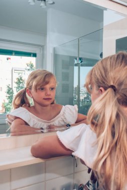 Banyoda iki uzun sarı kuyruklu sevimli sarışın çocuk aynaya bakıyor.