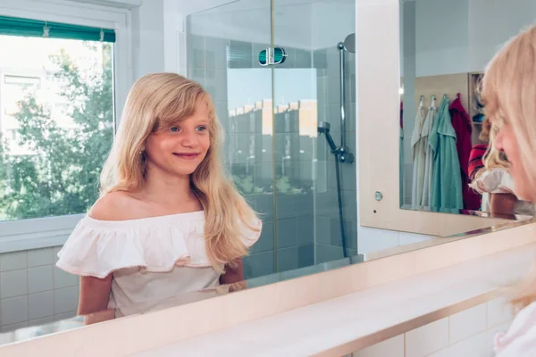 Encantadora chica rubia sonriendo en el baño delante del espejo —  Fotos de Stock