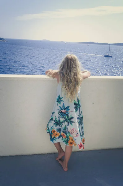 Menina adorável com cabelo loiro longo e vestido de verão olhando da varanda — Fotografia de Stock