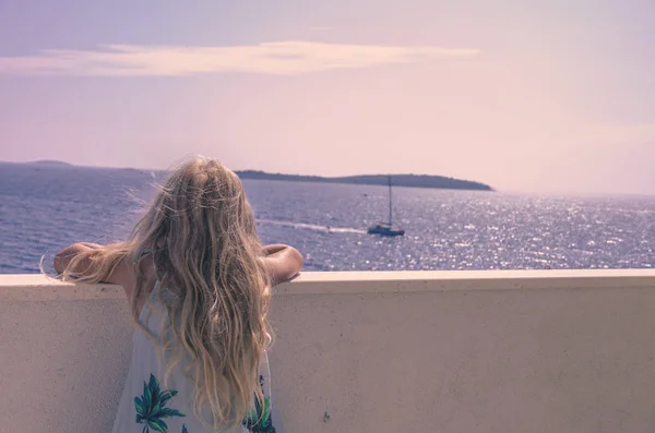 Menina adorável com cabelo loiro longo e vestido de verão olhando da varanda — Fotografia de Stock