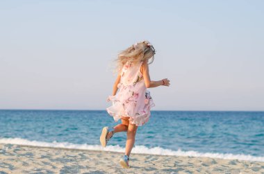 Uzun sarı saçlı, romantik elbiseli sevimli çocuk deniz kenarında tek başına dans ediyor.