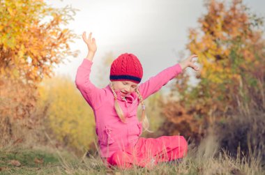 Güneşli bir sonbahar öğleden sonrasında çimenlerde oturan kırmızı şapkalı, tatlı, güler yüzlü kız.