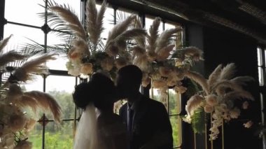 Gelin ve damat büyük bir pencere ve çiçek bir kemer arka planda öpücük.