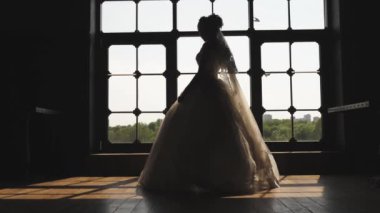 Gelinlikli gelin büyük pencerede dönüyor. Işık odaya pencereden girer. Gelinin elinde bir buket.
